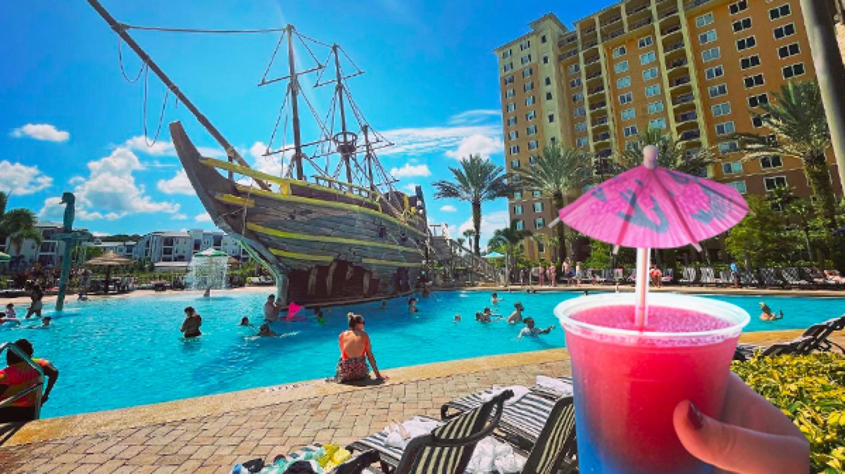 A frozen cocktail with a pink umbrella in the foreground, with the pirate ship pool behind
